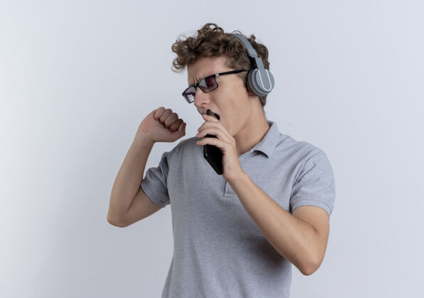 耳机戴着黑眼镜的年轻人身穿灰色马球衫 头戴耳机 手持智能手机 站在白墙上欣赏着他喜爱的音乐戴着抱着眼镜