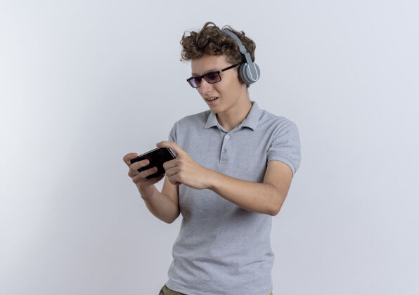 男人身穿灰色马球衫 头戴耳机 手持智能手机玩游戏的年轻人站在白墙上马球游戏站着