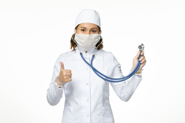 女性正面图年轻女医生戴无菌口罩因冠状病毒手持听诊器在白色表面病毒正面灭菌