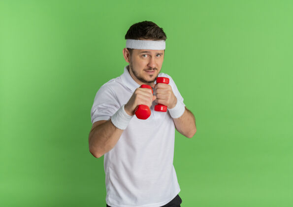 健身身穿白衬衫 头箍戴着哑铃 紧张地站在绿色背景下锻炼身体的年轻人站立哑铃紧张