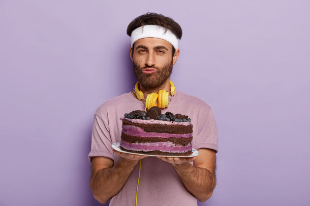 水平帅哥的画像没刮胡子的男人拿着美味的蛋糕放在盘子里 穿着休闲服 有着很好的意志不吃甜点靠着紫墙站着 享受着很好的味道男人用甜食站着猪鬃美味