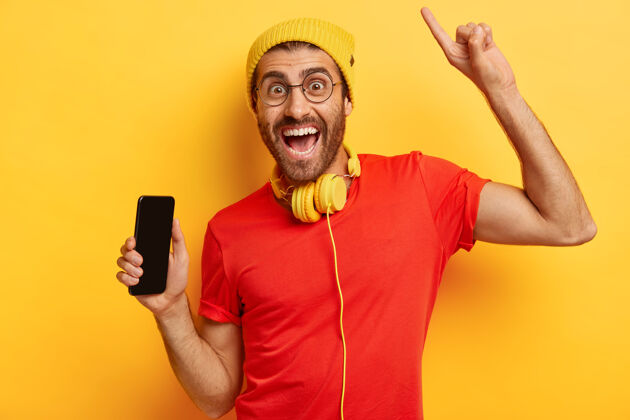 电话快乐无胡子男人的画像拿着空屏幕的手机 举起手臂并用食指指着上方 有着愉快的表情 戴着黄色帽子和红色t恤 戴着耳机移动短信黄色