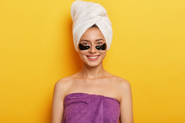 年轻快乐的欧洲女人 面带温和的微笑 有黑色的胶原蛋白斑块 减少黑眼圈的问题 用毛巾包在头上和身上 改善皮肤状况姿势女性面膜