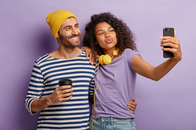 关系积极多样的情侣合影自拍 微笑扮鬼脸 喝外卖咖啡 穿休闲服 靠着紫色的墙拥抱科技 生活方式男人女性手机