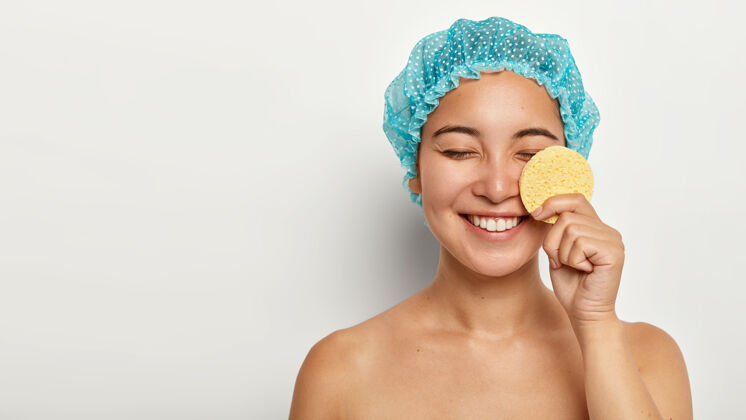 保持快乐的亚洲女人用化妆海绵洗脸 卸妆 闭上眼睛 戴上保护性的淋浴帽健康请特写