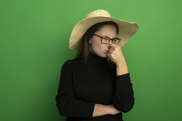 夏天年轻漂亮的女人戴着夏天的帽子 穿着黑色高领毛衣 戴着眼镜 闭着鼻子 脸上带着恼怒的表情翻过绿墙高领毛衣人帽子