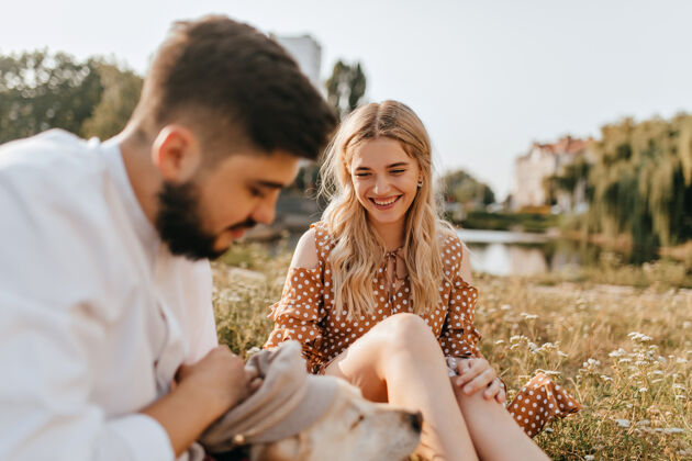 圆点连衣裙美丽的金发女孩和她善良的丈夫正在草地上休息 和他们的宠物玩耍这对夫妇在湖面的背景下摆姿势户外休闲微笑