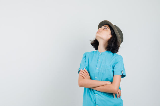积极年轻的女士交叉着双臂站着 看着天花板 穿着蓝色衬衫 戴着帽子女性女人喜悦