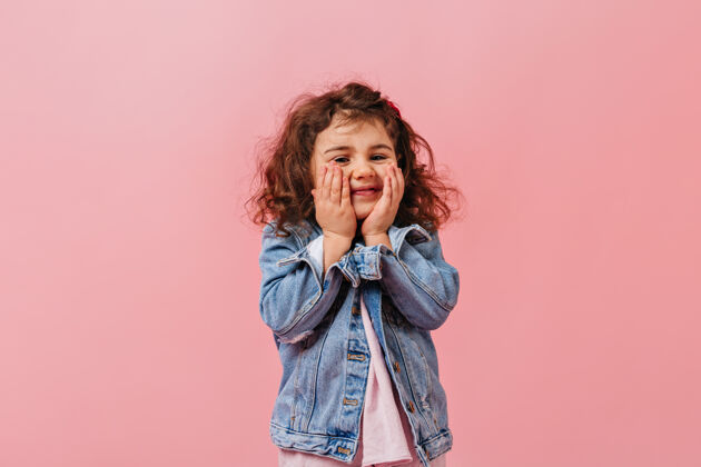 游戏可爱可爱的孩子在牛仔夹克触摸脸微笑的少女在粉红色的背景摆姿势学龄前儿童时尚表情