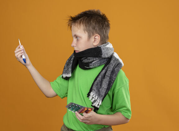 小的生病的小男孩穿着绿色t恤 脖子上围着暖和的围巾 手里拿着药片 站在橘色的墙上 看着他们困惑的样子男孩看着橘子