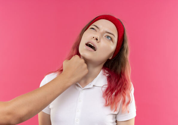 女人戴着头巾的年轻的运动型女人用拳头顶着粉色的墙 下巴上打了个巴掌运动员运动装市民