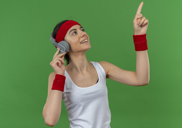 健身戴着头带和耳机 穿着运动服的年轻女健身者微笑着 用食指指着绿色的墙壁微笑耳机站立