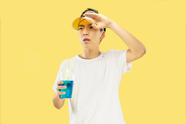 情绪黄色工作室背景上的韩国年轻人半身像穿着白衬衫 戴着黄色帽子的男模喝鸡尾酒人类情感的概念 表达 夏天 假期 周末霓虹灯喝酒太阳镜