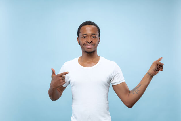 男人在蓝色背景上 一个穿着白衬衫的非洲裔美国年轻人的半身特写肖像人类的情感 面部表情 广告概念展示空酒吧 指向 选择 邀请商人成功顺序