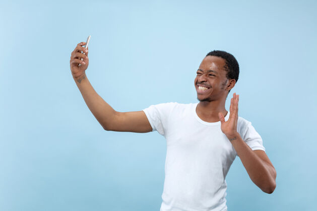 公司半身特写 蓝色背景 身穿白衬衫的非洲裔美国年轻男子人类情感 面部表情 广告概念为社交媒体制作自拍或内容 虚拟博客胡须媒体顺序