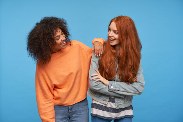 20多岁室内拍摄的迷人的积极的女朋友欢笑 而看着对方 在良好的心情 而站在蓝色的墙壁外套非洲女性