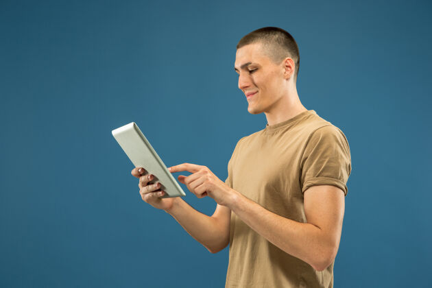 面部在蓝色工作室背景上的白人年轻人半身肖像穿着衬衫的漂亮男模人类情感的概念 面部表情 销售 广告使用平板电脑进行自拍 视频日志或学习表情人成功