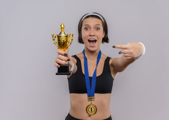 手指一位身穿运动服 脖子上挂着金牌的年轻健身女士拿着她的奖杯 用手指着奖杯 高兴而兴奋地站在白墙上为自己的成功而欢呼人物快乐兴奋