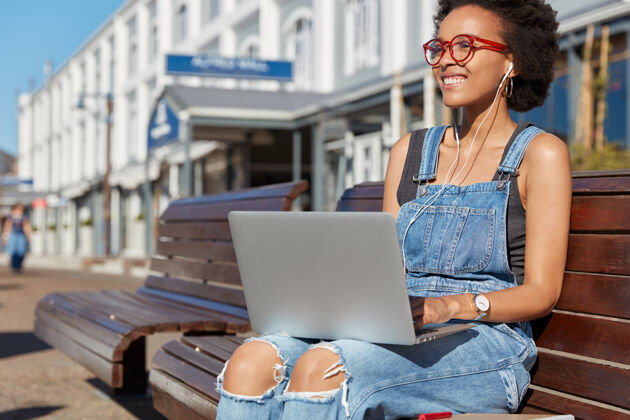 街道积极向上的美国黑人妇女戴着耳机听音乐 在笔记本电脑上工作 穿着牛仔工作服 面带微笑 坐在城市环境的长凳上 专注于远方笔记本阅读工作服