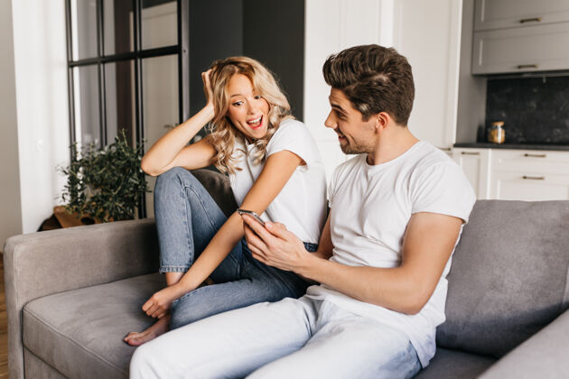 手机一对年轻的情侣坐在沙发上兴奋的男人用手机向他的女朋友展示新闻惊讶的女孩看着手机屏幕 开心地笑着沙发室内关系
