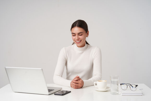 微笑年轻可爱的黑发女士 面容自然 双手合十 坐在白色墙壁上的桌子旁 面带微笑 穿着正式的衣服年轻手机模特