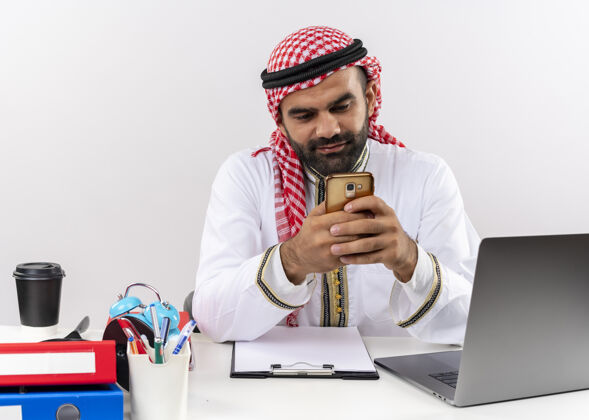 笔记本电脑身着传统服装的阿拉伯商人坐在桌旁 手提电脑拿着智能手机在办公室发短信信息坐着电脑