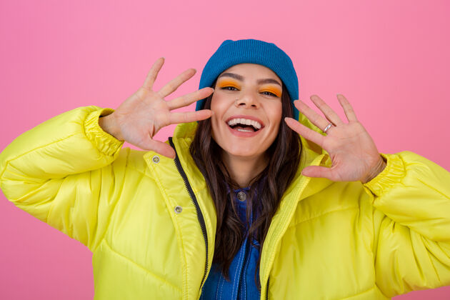 运动装迷人微笑的时尚女性的肖像在冬天的时尚造型在粉红色的墙上穿着鲜艳的霓虹黄夹克 戴着蓝色的针织帽子 穿着暖和的衣服运动快乐衣服