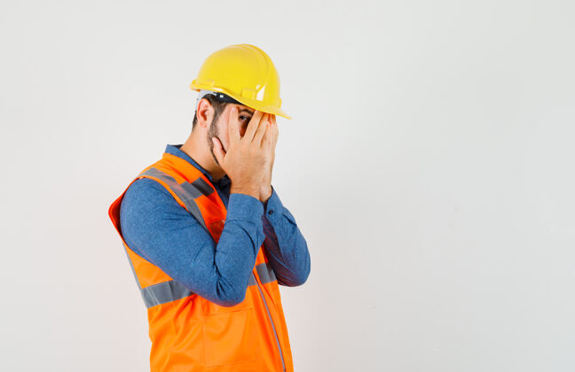 安全年轻的建筑工人穿着衬衫 背心 头盔透过手指看 看起来很害怕 前视图制服专业工具