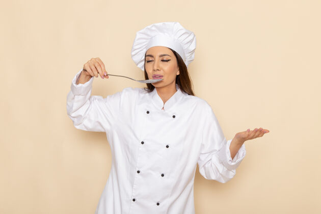 年轻身穿白色厨师服的年轻女厨师手持大银勺在白墙上品尝美食的正面图女大人