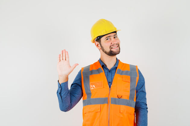 建筑年轻的建筑工人穿着衬衫 背心 头盔挥手致意或告别 看上去很高兴建筑工人工程制服