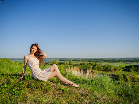 顶坐在悬崖边的年轻女子穿着白色连衣裙的漂亮女孩在户外摆姿势女模特在田野里摆姿势一个阳光明媚的夏日放松春天腿
