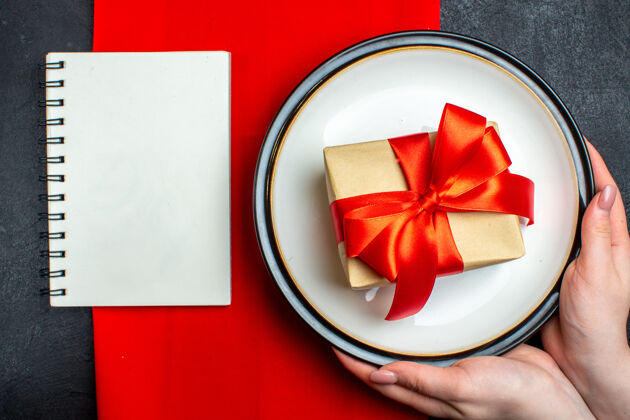 空盘子国家圣诞餐背景俯视图 手拿着空盘子 蝴蝶结形状的红丝带 黑色桌子上的红餐巾上放着一个笔记本膳食国家笔记本