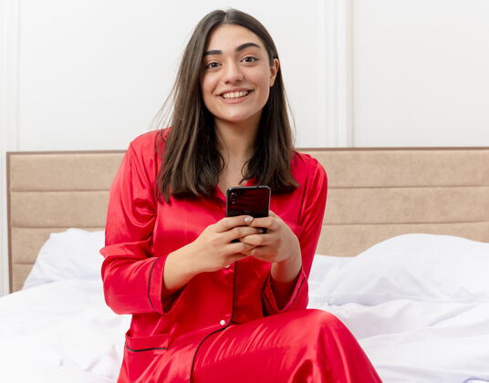 床穿着红色睡衣的年轻美女坐在床上 用智能手机微笑着看着卧室里的摄像头 背景光线很亮室内女人漂亮