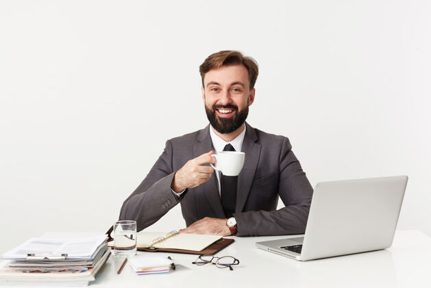 杯子快乐的年轻留着棕色短发的商人在现代办公室工作 在开会前喝一杯咖啡 坐在白色的墙壁上开心地微笑着男士西装积极