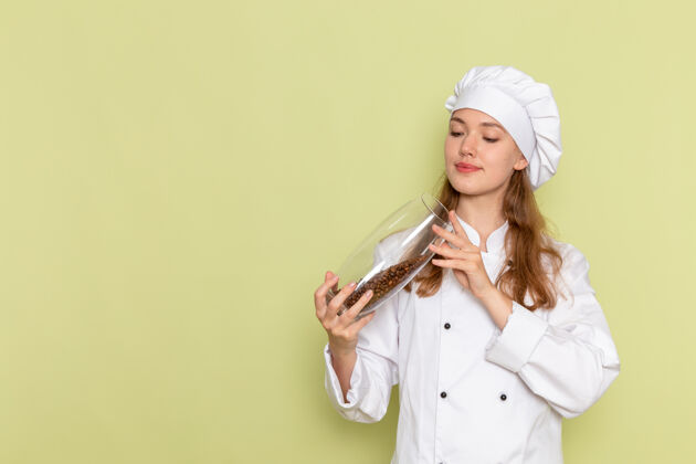 举行正面图：女厨师穿着白色厨师服拿着装满咖啡籽的罐子放在绿色桌子上 厨房里的饭菜是女性的颜色厨房厨师套装