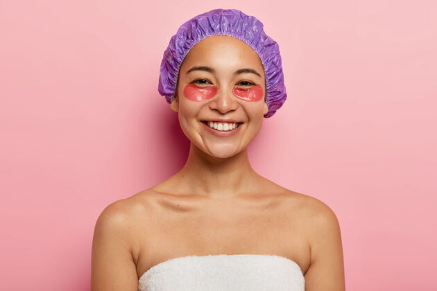 表情美容护肤理念开朗的亚洲女性微笑积极 在眼睛下贴水凝胶贴片 戴淋浴帽 赤膊上阵 裹着白毛巾 隔离在粉红色的墙上面部贴片裸露