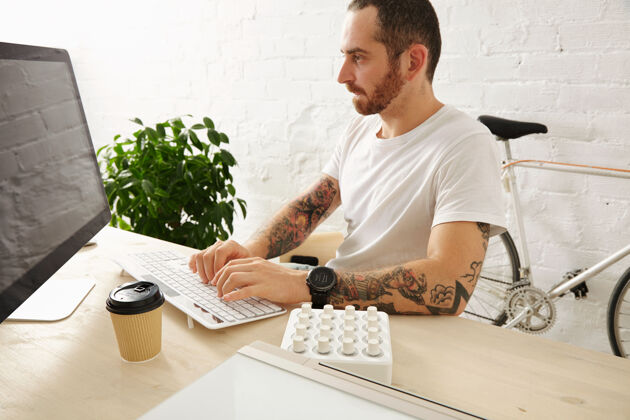 计算机留着胡子的纹身男子穿着白色t恤在家里的电脑上工作 侧视图 夏日时光打字看桌子