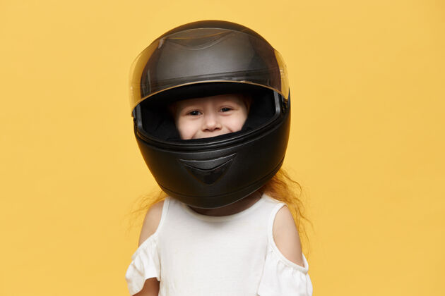 摩托车手可爱顽皮的小女孩戴着从她父亲那里拿来的黑色摩托车头盔滑稽的女孩儿在保护马达设备里摆出孤立的姿势 面带微笑摩托车手骑手保护