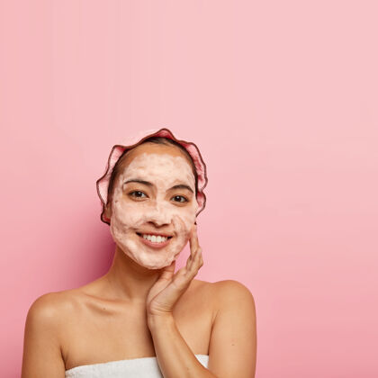 女人年轻的中国女人喜欢净化面部皮肤 用肥皂洗 摸脸颊 微笑着看 清洁毛孔 戴浴帽 在室内摆姿势 在粉红色的墙上复制空间皮肤年轻应用
