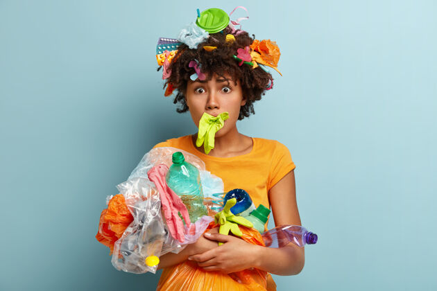 女人尴尬的年轻卷曲的美国黑人妇女的照片嘴里戴着橡胶手套 携带塑料垃圾 担心全球环境污染 隔离在蓝色的墙上生态概念不安垃圾瓶子