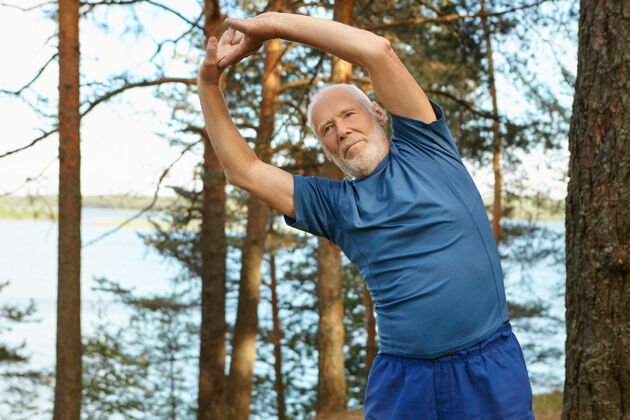 运动装精力充沛 自强不息的大胡子老人穿着运动服 在户外与森林和河流合影 双臂高举 侧弯 跑步前热身锻炼锻炼成熟男人