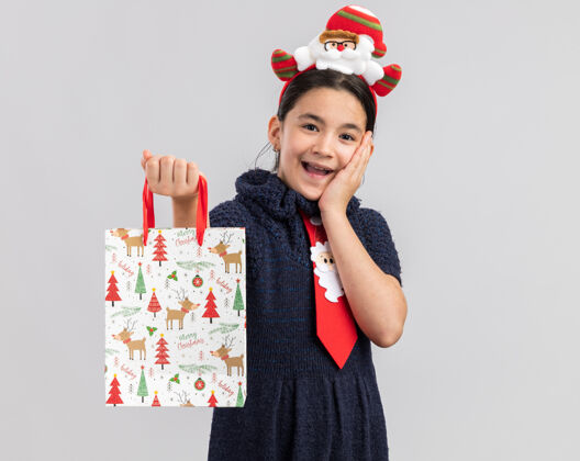 包小女孩穿着针织连衣裙 头上系着一条红色领带 头上戴着一个有趣的圣诞边 手里拿着一个装有圣诞礼物的纸袋 看上去很惊讶头编织抱着