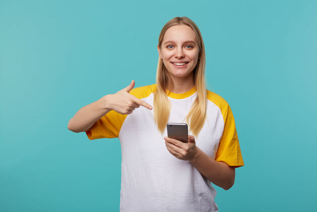 高兴年轻的白头女人的肖像在她的智能手机上愉快地展示着她举起的手和迷人的微笑 摆出蓝色的姿势情绪黄色情绪