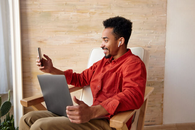 自由职业者快乐的年轻黑发黑皮肤男性 留着胡须 一边愉快地微笑 一边打视频电话 一边用笔记本电脑在家里远程工作笔记本电脑遥控电话