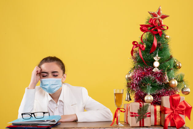 医生正面图女医生戴着防护面具坐在黄色背景上 戴着圣诞树和礼品盒病毒正面背景