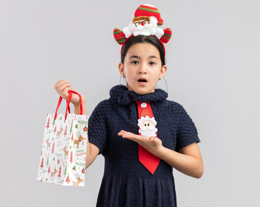 快乐穿着针织连衣裙的小女孩头上系着一条红色领带 头上戴着一个有趣的圣诞边 手里拿着一个纸袋 手里拿着圣诞礼物 一副迷茫的样子站看女孩