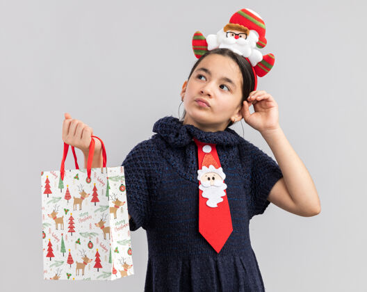 圣诞快乐穿着针织连衣裙的小女孩头上系着一条红色领带 头上戴着一个滑稽的圣诞边 手里拿着一个装有圣诞礼物的纸袋 困惑地看着一边领带困惑包