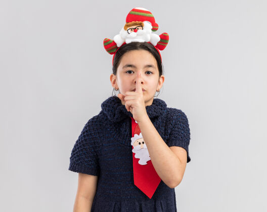 圣诞快乐小女孩穿着针织连衣裙 头上系着一条红色领带 戴着一个有趣的圣诞戒指 手指放在嘴唇上 做着沉默的手势制作编织看