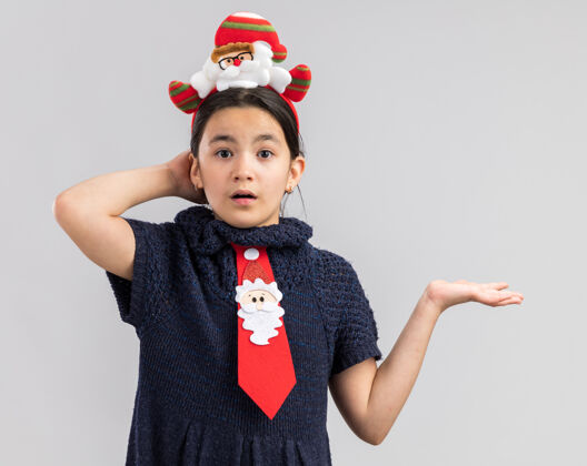 新小女孩穿着针织连衣裙 头上系着一条红色领带 头上戴着一个滑稽的圣诞边 看起来很困惑 胳膊伸出来了快乐领带裙子