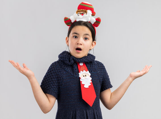 圣诞快乐小女孩穿着针织连衣裙 头上系着一条红色领带 头上戴着一个滑稽的圣诞边 双臂高举 看起来很惊讶圣诞节头巾连衣裙